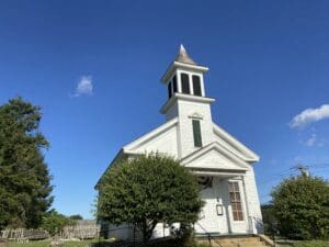 A white church with a steeple in the middle of a grassy field.